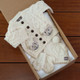 Luxury Aran Baby Gift Set. A closed, light brown gift box contains a knitted cream-colored baby outfit. The ensemble includes a buttoned Aran cardigan featuring sheep motifs, a cozy hat, and matching booties with cable knit patterns. The box is placed on a wooden surface. Luxury Aran Baby Gift Set. A closed, light brown gift box contains a knitted cream-colored baby outfit. The ensemble includes a buttoned Aran cardigan featuring sheep motifs, a cozy hat, and matching booties with cable knit patterns. The box is placed on a wooden surface.